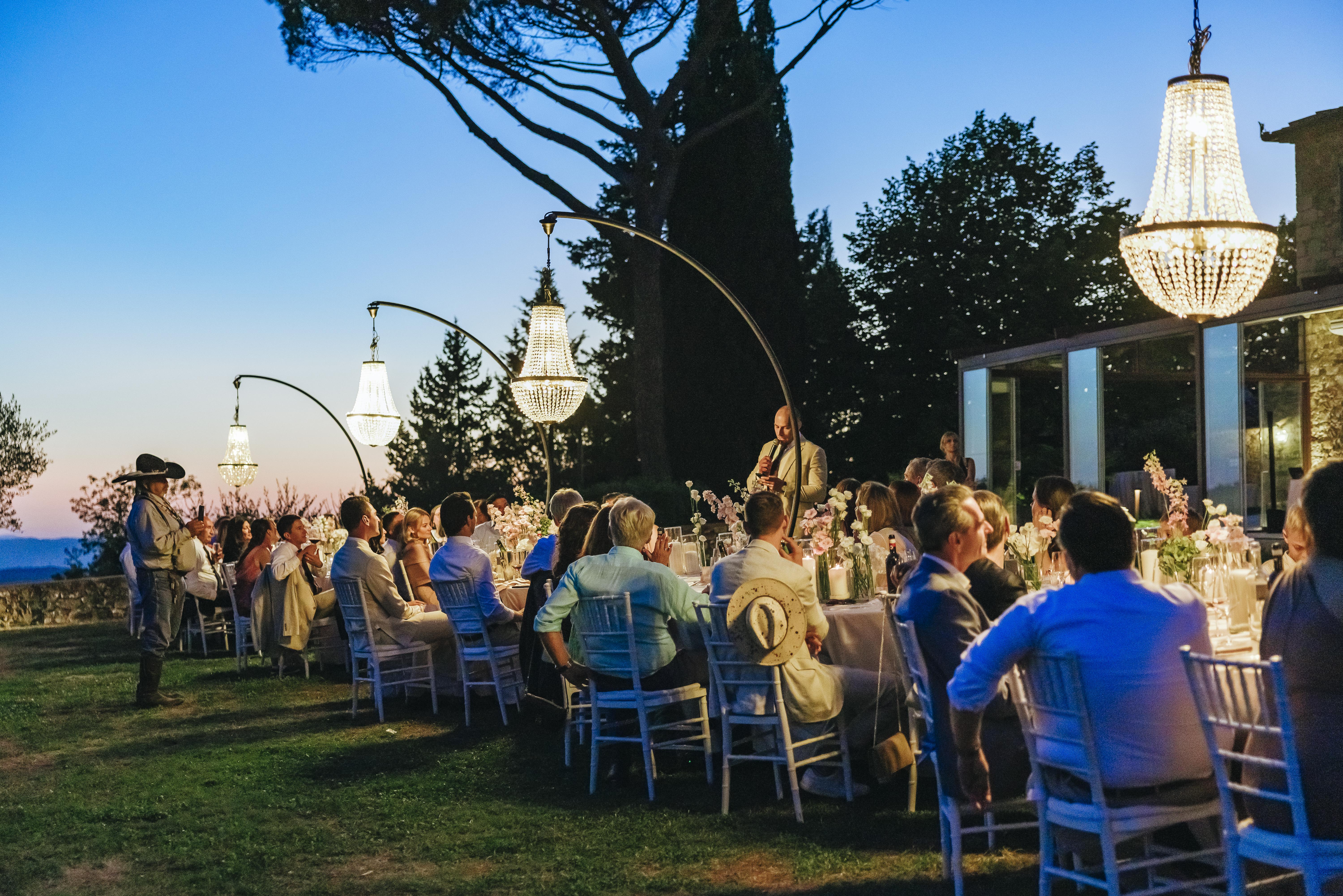 Wedding guests enjoying a candlelit dinner under chandeliers at dusk