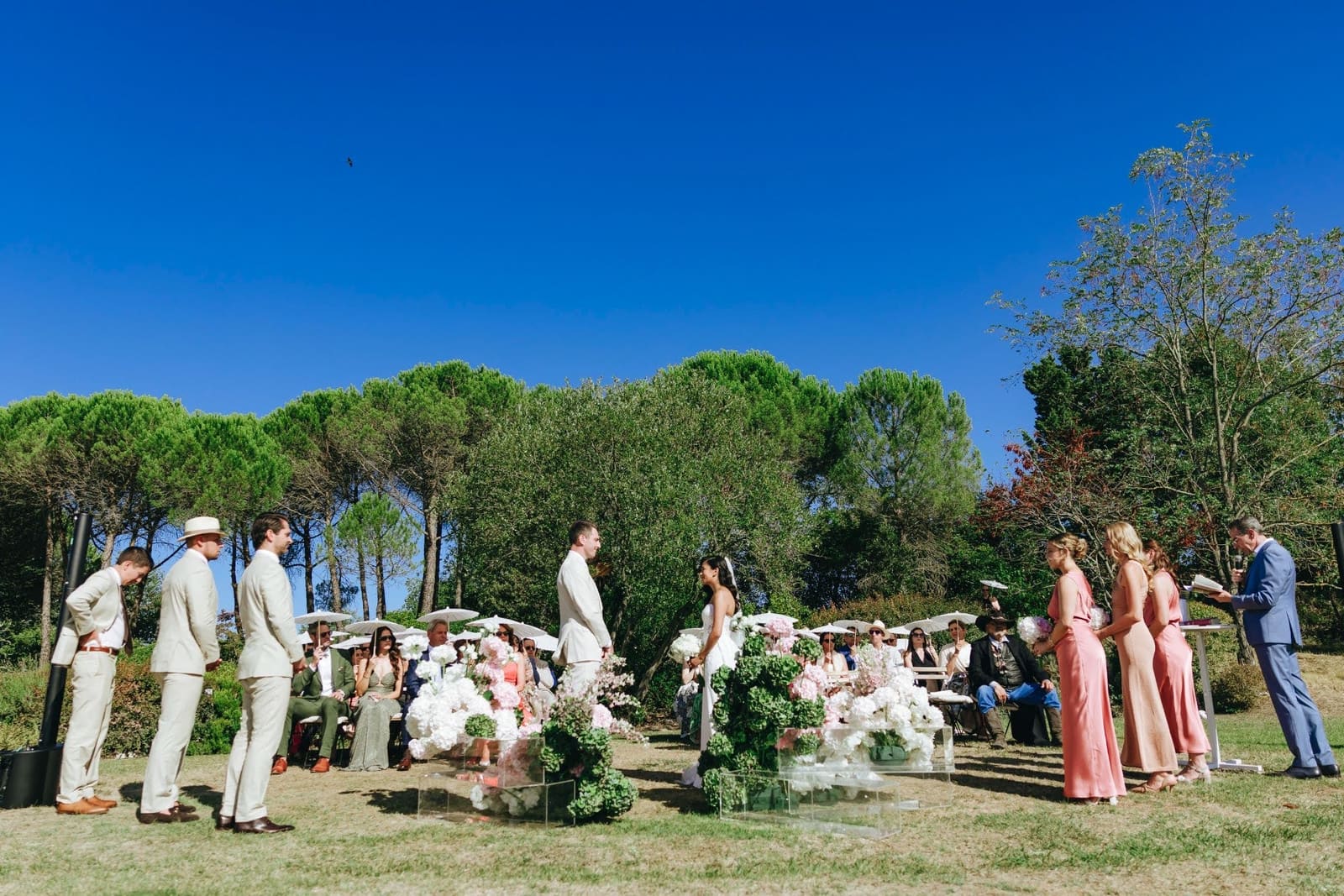Wedding guests seated with parasols on the lawn at Le Filigare in Tuscany, watching the ceremony under a clear sky