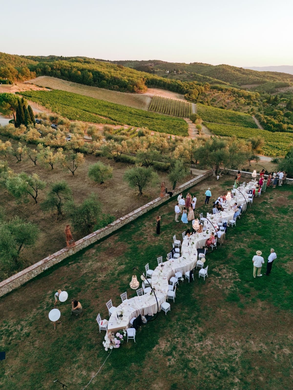 Aerial view of one long dinner table curving across the lawn at golden hour, guests standing beside it, vineyards and Tuscan hills behind